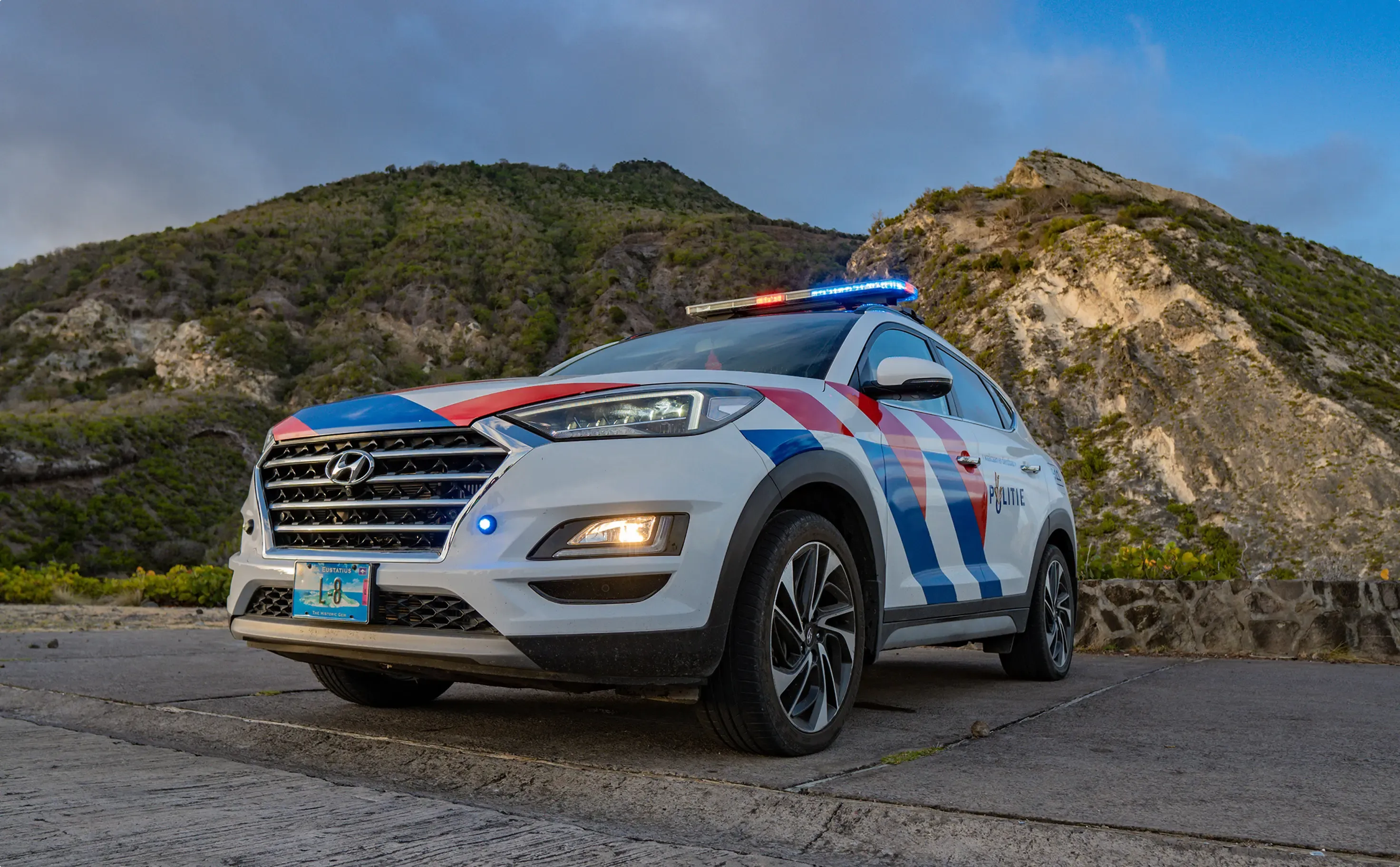 Police car of the Caribbean Netherlands Police Force with red and blue striping and emergency lights, photographed against the mountainous landscape of Saba Island.